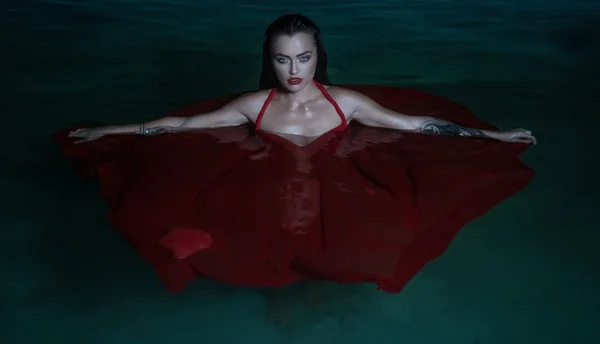 Красивая загадочная женщина, глядя, как вампир, носить красное платье, глядя в камеру ehile стоя в бассейне ночью — стоковое фото